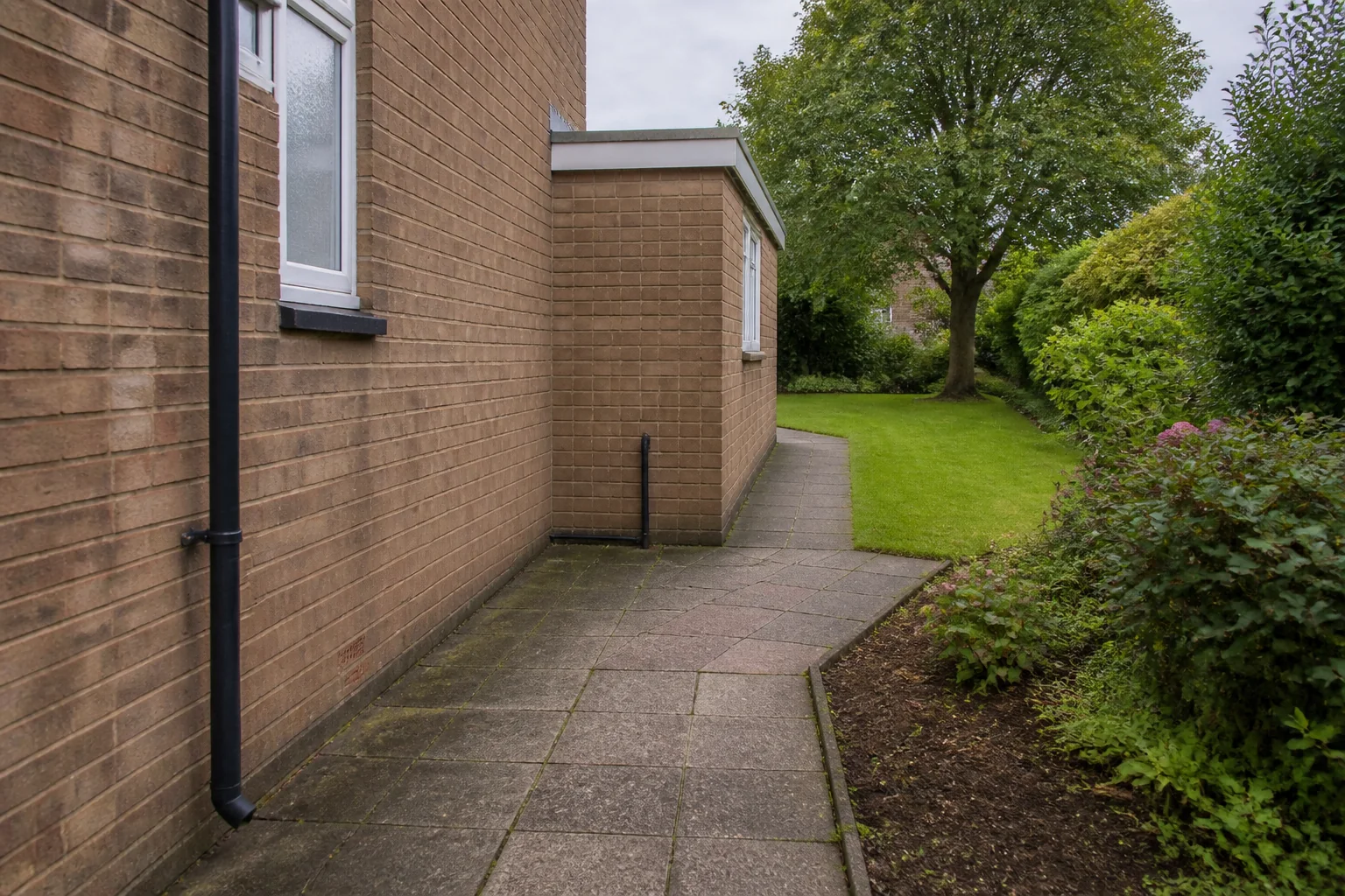 A worn slab path turns around a single-storey brick extension toward a lawn and mature broadleaf tree.