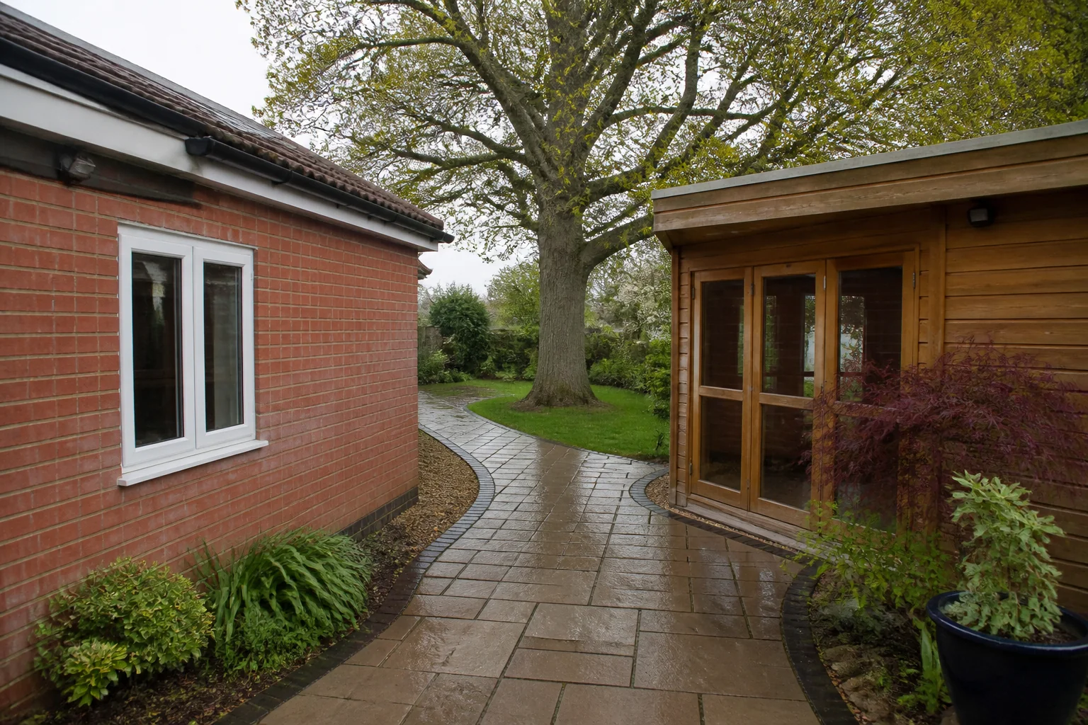 A paved route curves between a red-brick house extension and a timber garden room toward a mature tree.