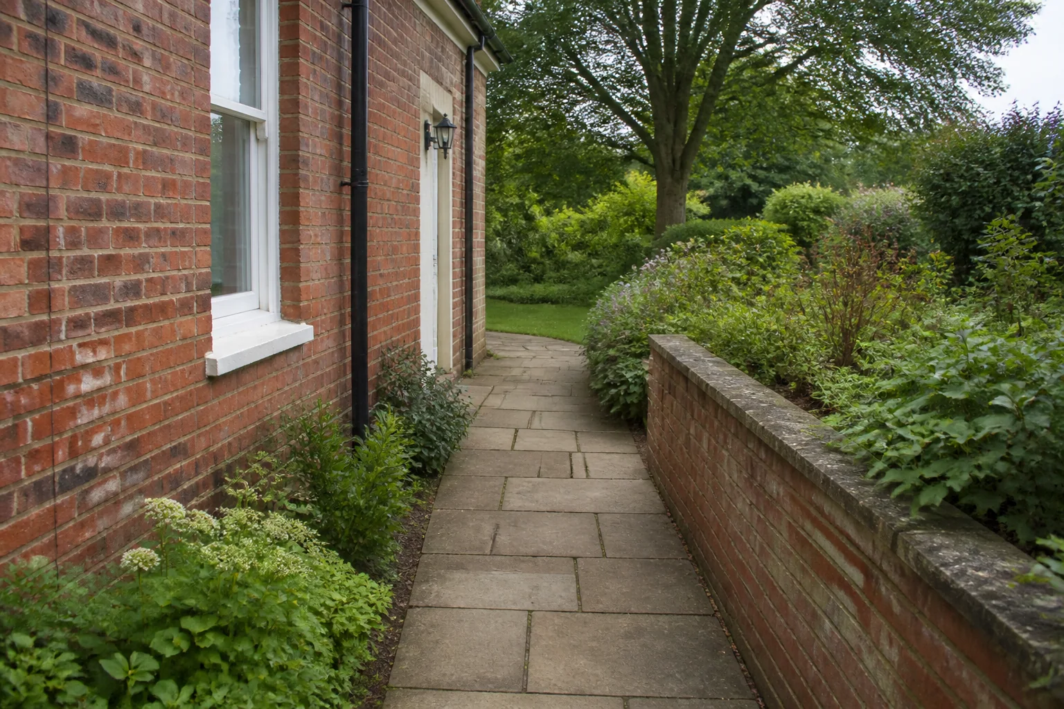 A paved route passes between an older red-brick house and a low planted wall before opening toward a mature tree.