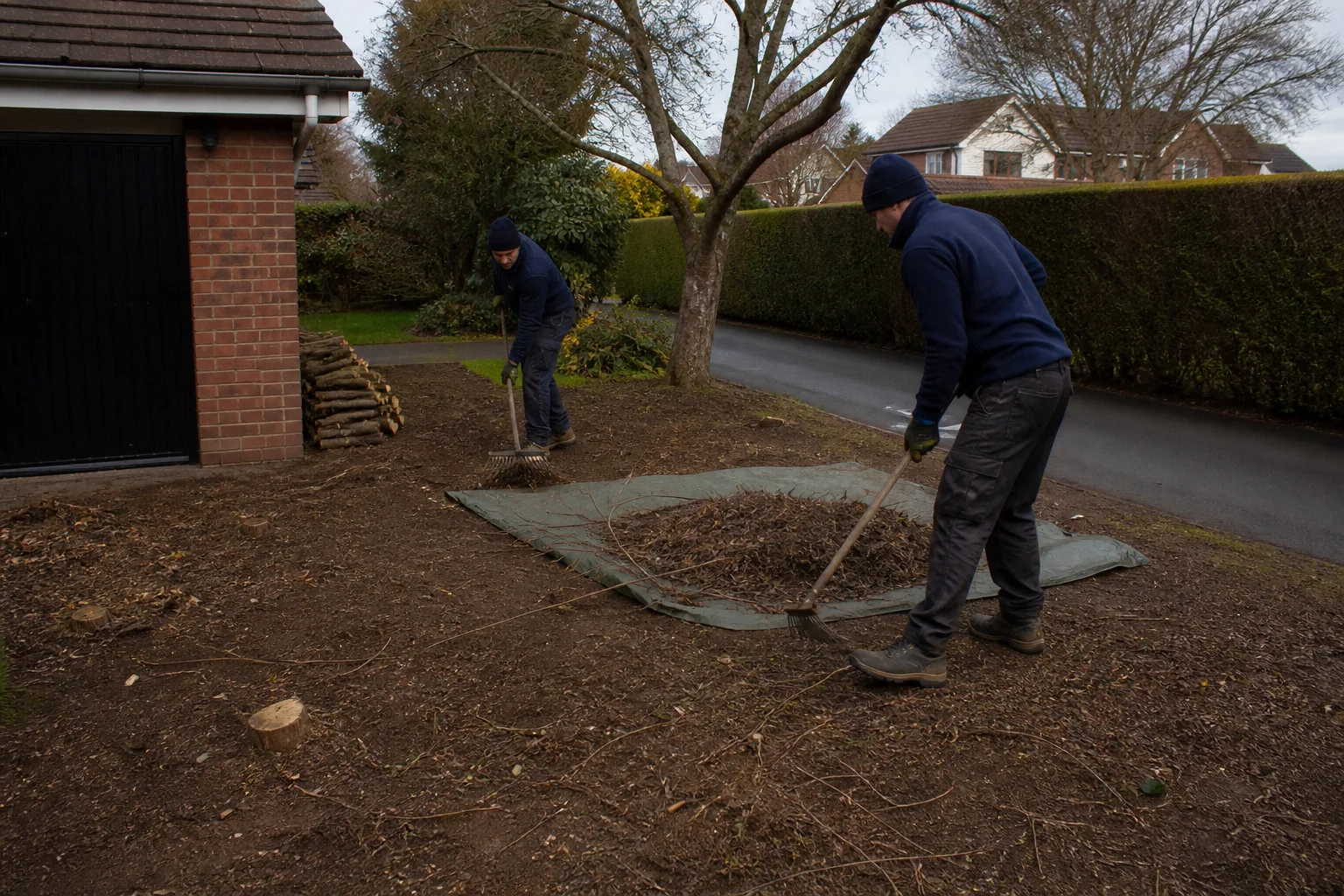 Two workers rake a cleared garden area with a mature tree and hedge retained.