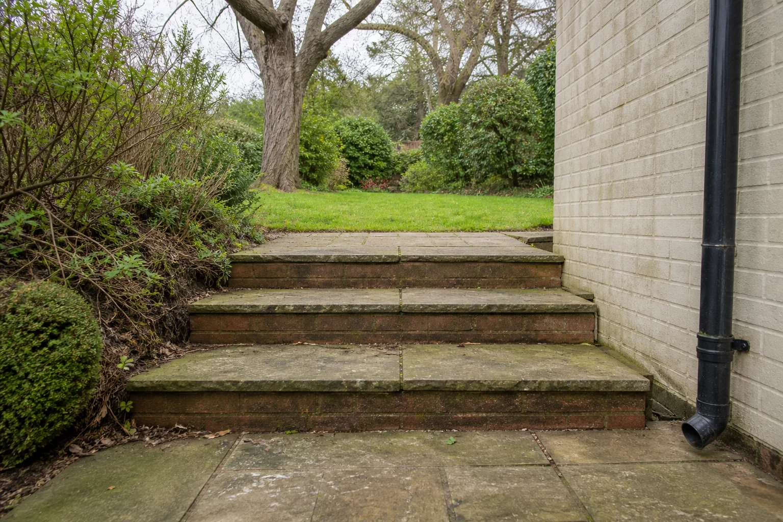 Three broad brick steps rise beside a pale brick house and planted bank to an upper lawn with mature trees.