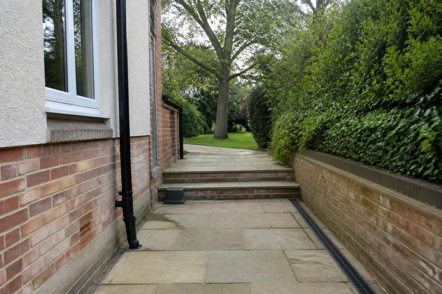 A paved side route rises by two shallow steps between a house wall and a planted boundary before reaching a mature tree.
