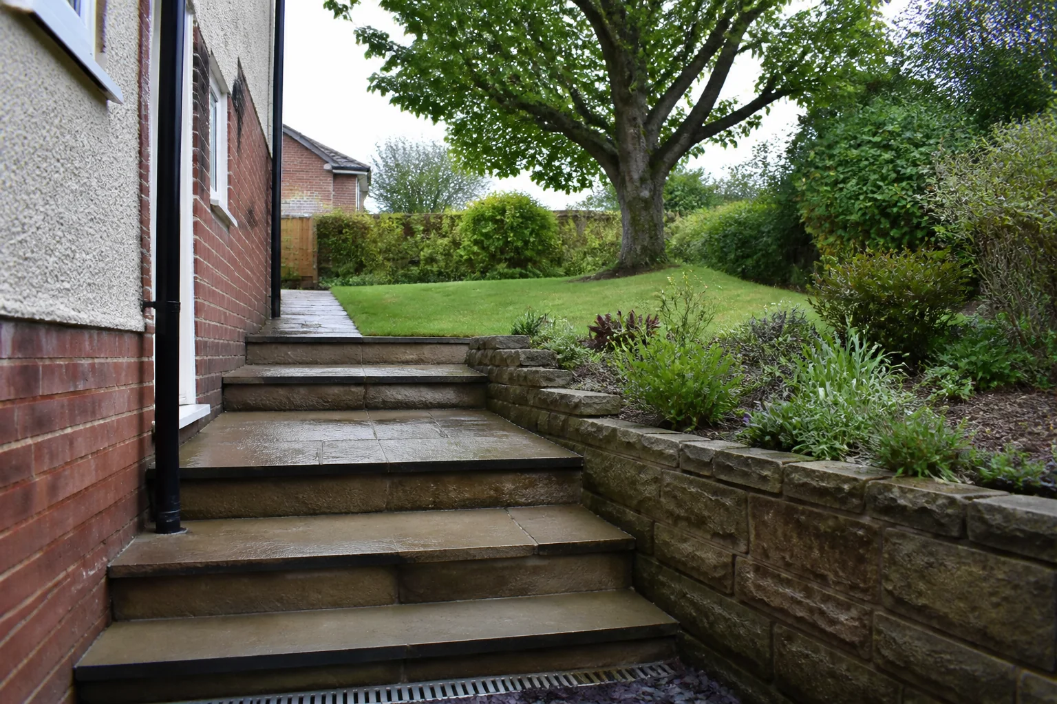 A paved garden route rises through broad stone steps beside a low retaining wall toward a mature tree on an upper lawn.