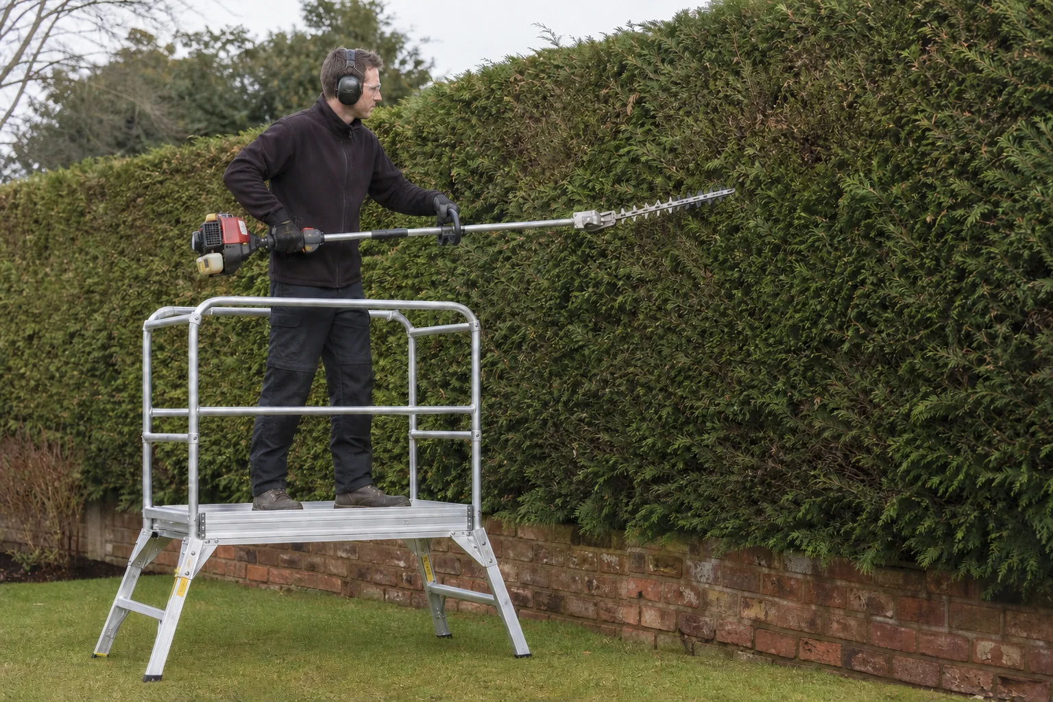 A tree worker uses a petrol long-reach hedge cutter from a guard-railed platform.