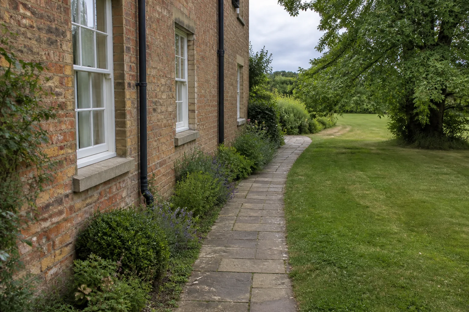 A long stone path bends beside an older brick house and planted edge toward a mature broadleaf tree on an open lawn.