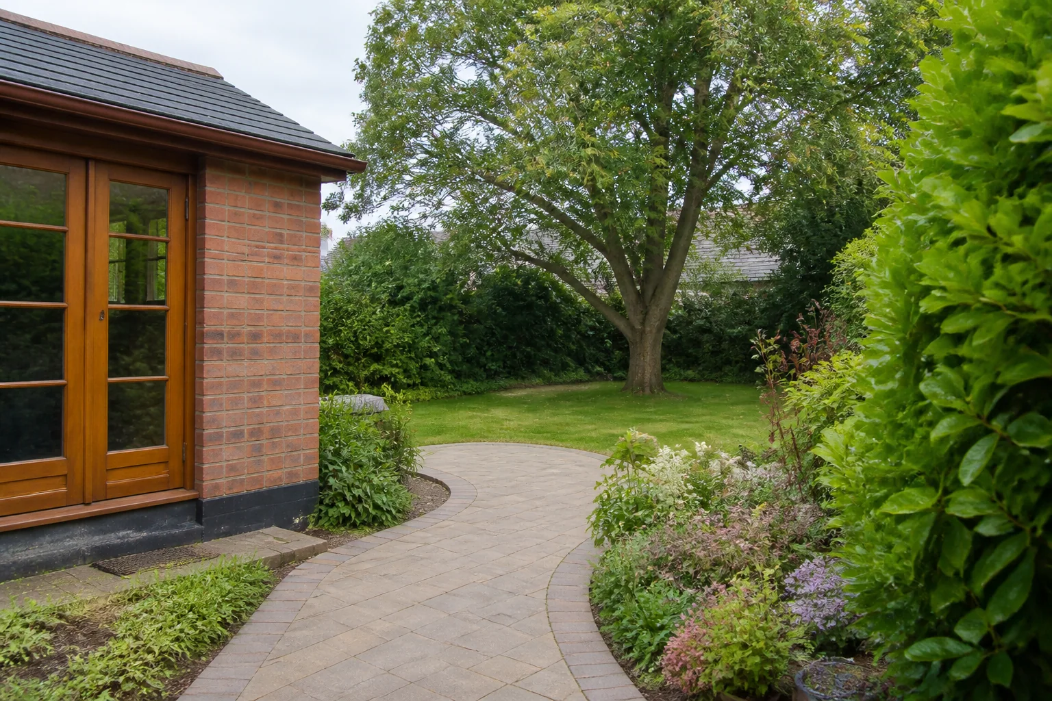 A curved paved garden path passing a modest brick garden room towards a mature broadleaf tree.