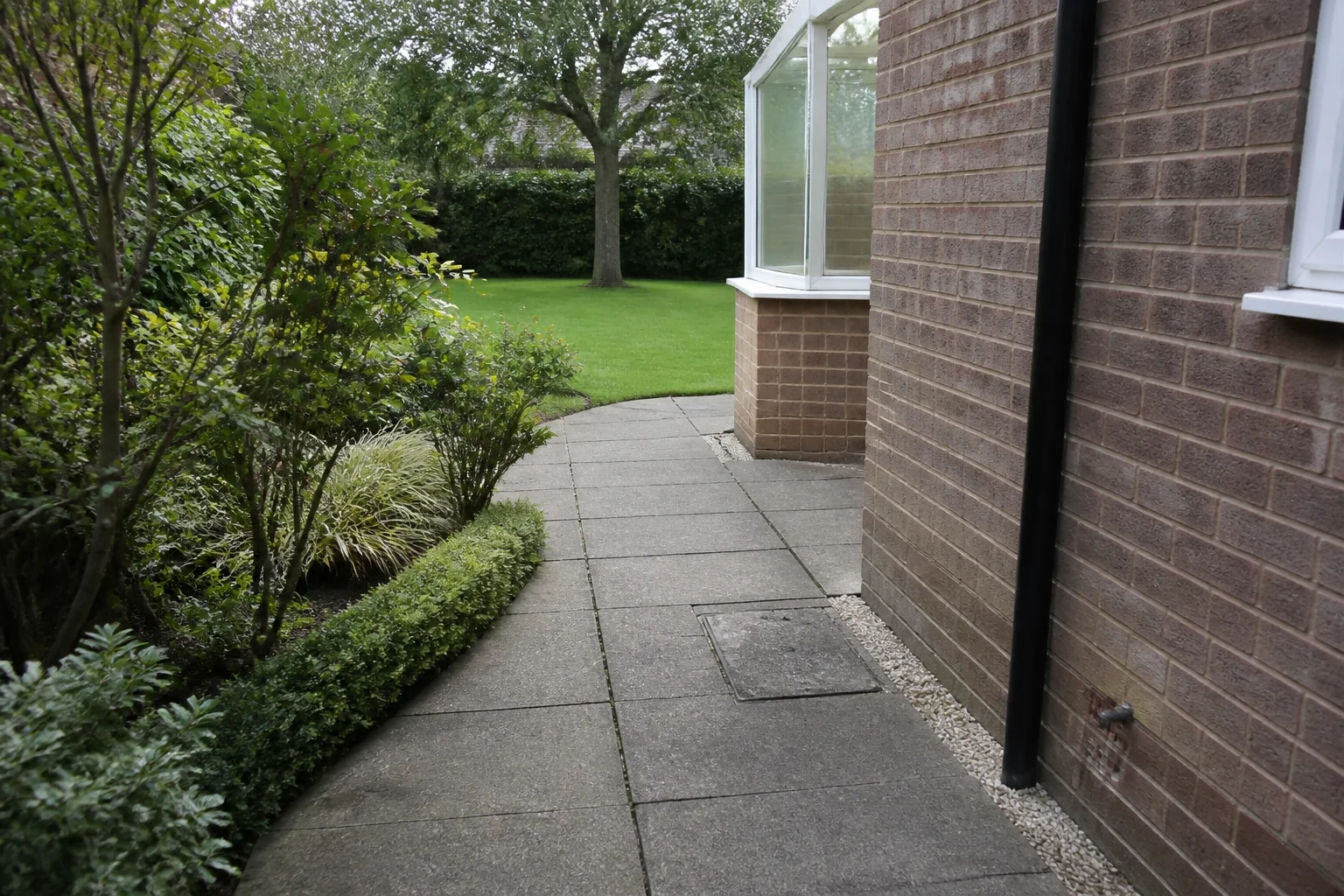 A slab path turns around a brick conservatory corner between a house wall and established planting toward a mature tree.