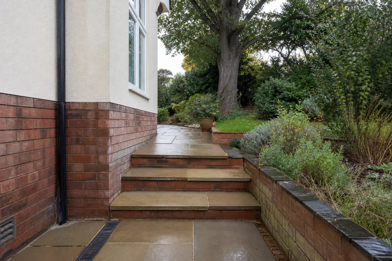 A paved route rises by broad shallow steps between a house wall and a planted retaining bed toward a mature tree.