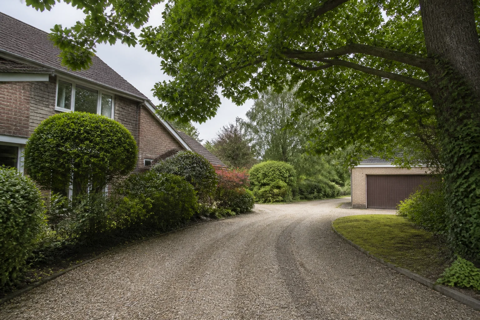 A long curved gravel drive passing a detached brick home and garage beneath mature broadleaf branches.