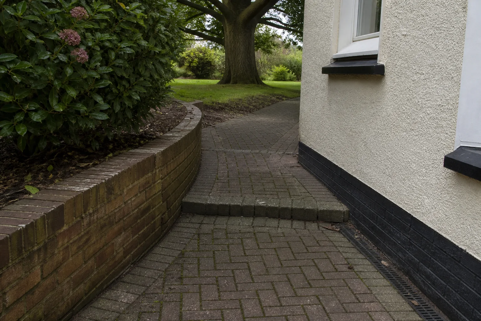 A block-paved route curves between a low brick retaining wall and a rendered house corner toward a mature tree.