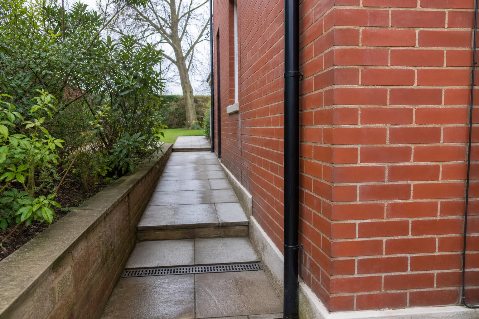A paved route with one shallow step between a red-brick house wall and a low planted bed, with a mature tree beyond.